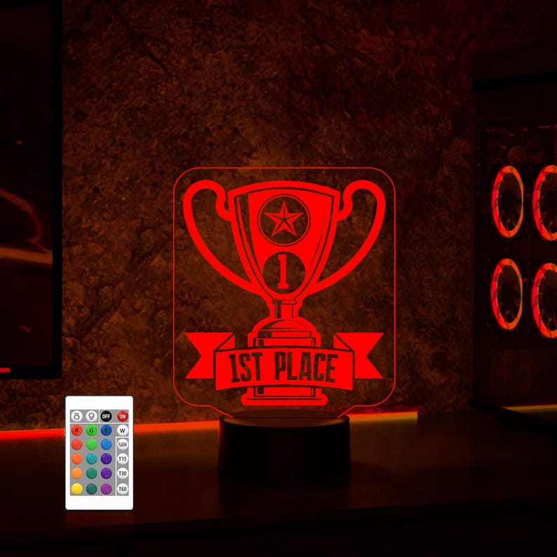 Puede incluir: Una luz nocturna LED roja con forma de trofeo con una pancarta que dice "1er lugar". La luz est&aacute; sobre una base negra y viene con un mando a distancia.