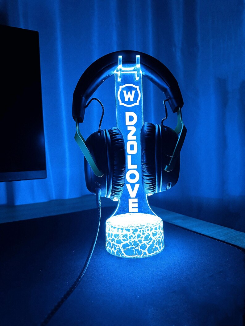 Op de afbeelding: Een blauwe LED-verlichte standaard met een gebarsten glazen basis, die een zwarte headset vasthoudt. De standaard heeft de letters "W D2OLOVE" erop gedrukt.