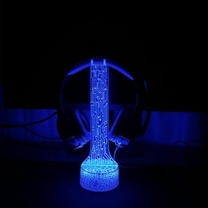 Puede incluir: Una luz LED azul con un dise&ntilde;o de placa de circuito, con forma de cilindro alto, con unos auriculares negros apoyados en la parte superior.