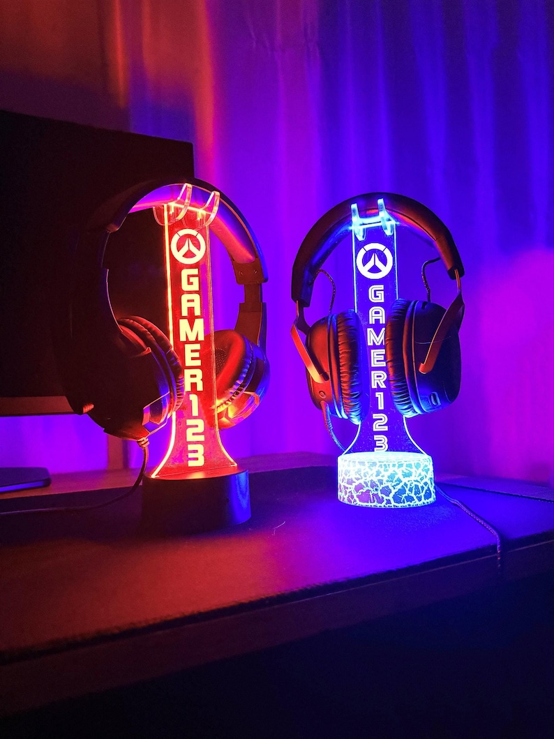 May include: Two gamer headset stands with a glowing red and blue light. The stands are made of clear acrylic and have the word "GAMER123" printed on them. The stands are lit from the bottom and have a white base.