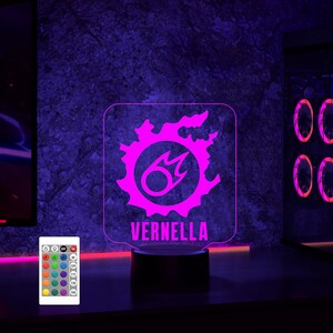 May include: A pink and white LED night light featuring a stylised fire symbol with the name "VERNELLA" printed below. The light sits on a black base and comes with a remote control offering various colour options.