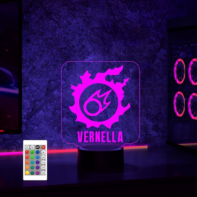 May include: A pink and white LED night light featuring a stylised fire symbol with the name "VERNELLA" printed below. The light sits on a black base and comes with a remote control offering various colour options.