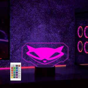 May include: A pink raccoon logo illuminated by a 3D LED lamp with a remote control. The lamp has a black base and is on a dark surface.