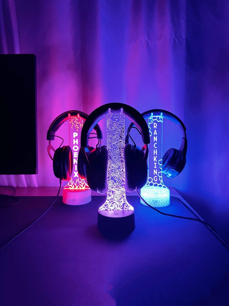 May include: Three illuminated headphone stands with headphones. The stands have a black base and a clear acrylic top with a design. The stand on the left says "PHOENIX" in red, the middle stand has a topographic design, and the right stand says "RANCHKING" in blue.