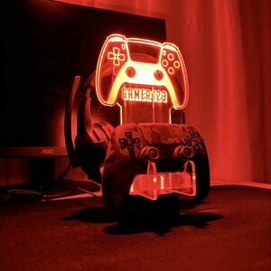 Op de afbeelding: Een rood verlichte gaming controller stand met de tekst "GAMER 123". Een camouflage patroon controller rust op de standaard, met een headset erachter. De standaard wordt verlicht door een rode gloed.