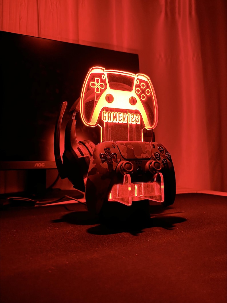 Op de afbeelding: Een rood verlichte gaming controller stand met de tekst "GAMER 123". Een camouflage patroon controller rust op de standaard, met een headset erachter. De standaard wordt verlicht door een rode gloed.