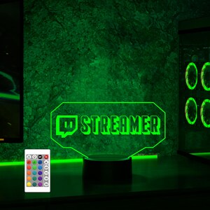 Op de afbeelding: Een groen LED-licht met een witte basis en een afstandsbediening. Het licht heeft een Discord-logo en het woord "STREAMER".