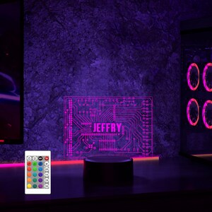 K&ouml;nnte beinhalten: Ein lila und pinkfarbenes 3D-gedrucktes Nachtlicht mit einem Leiterplatten-Design und dem Namen "JEFFRY" in der Mitte. Das Licht steht auf einem schwarzen Sockel und wird mit einem rosa Schein beleuchtet. Eine Fernbedienung mit mehreren Farboptionen ist im Lieferumfang enthalten.