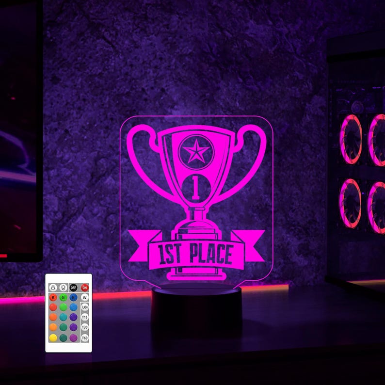 Puede incluir: Una luz nocturna rosa impresa en 3D con forma de trofeo con una pancarta que dice "1er lugar". La luz nocturna est&aacute; sobre una base negra y tiene un control remoto con m&uacute;ltiples opciones de color.