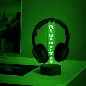 Puede incluir: Una luz LED verde con una base negra y una inscripci&oacute;n blanca que dice "GamerTag". La luz est&aacute; iluminada y tiene unos auriculares negros apoyados en la parte superior.