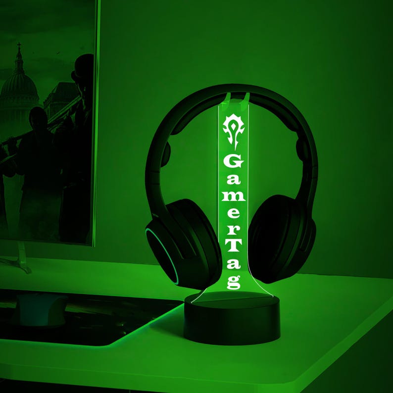 Puede incluir: Una luz LED verde con una base negra y una inscripci&oacute;n blanca que dice "GamerTag". La luz est&aacute; iluminada y tiene unos auriculares negros apoyados en la parte superior.
