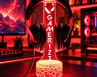 Könnte beinhalten: Ein schwarzes kabelgebundenes Headset auf einem vertikalen Acrylständer. Der Ständer hat eine leuchtend rote Basis mit Rissstruktur und eine transparente aufrechte Platte mit dem weißen Text GAMER1ZM und einem Logo.