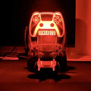 Op de afbeelding: Een rood verlicht LED-licht in de vorm van een gamecontroller, met de tekst "GAMER 123". Een andere controller is hieronder zichtbaar, rustend op een standaard. De afbeelding is tegen een donkere achtergrond geplaatst, wat een dramatisch effect cre&euml;ert.