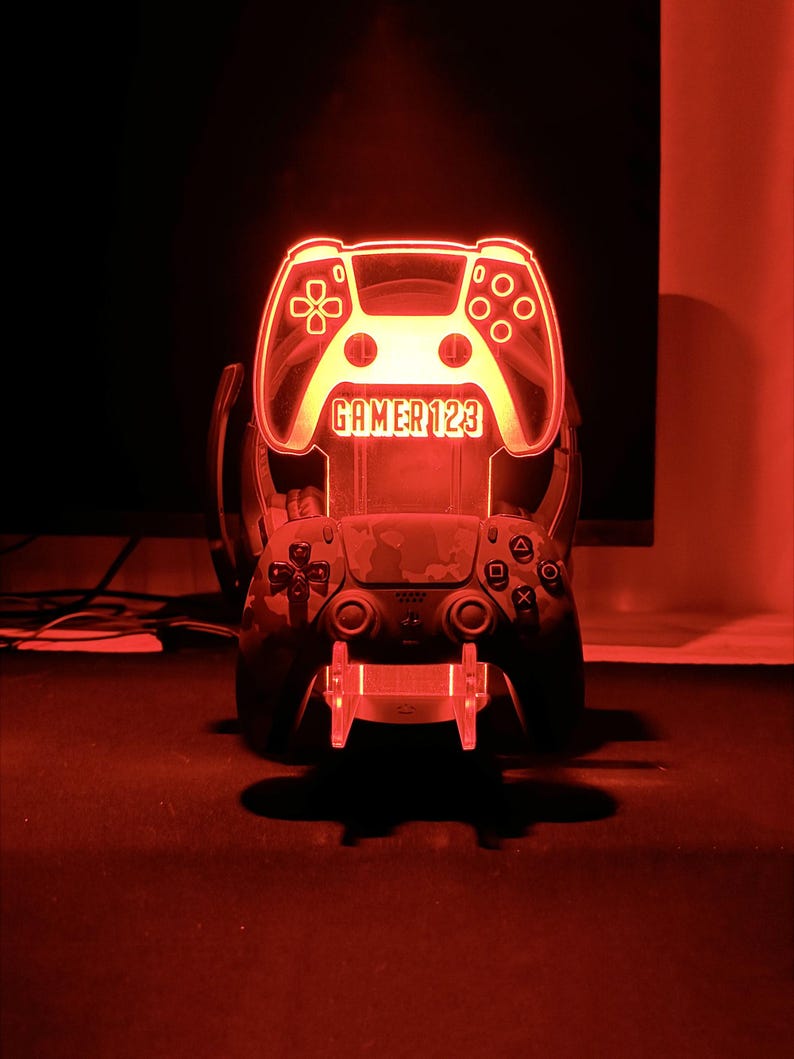 Op de afbeelding: Een rood verlicht LED-licht in de vorm van een gamecontroller, met de tekst "GAMER 123". Een andere controller is hieronder zichtbaar, rustend op een standaard. De afbeelding is tegen een donkere achtergrond geplaatst, wat een dramatisch effect cre&euml;ert.