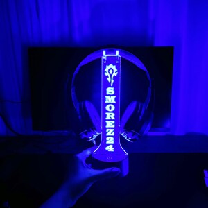 Peut inclure: Un support de casque bleu illumin&eacute; avec le texte "SMORENZ24" et un logo blanc. Le support est con&ccedil;u pour maintenir un casque et est &eacute;clair&eacute; d'une lueur bleue. Le support est tenu par une main.