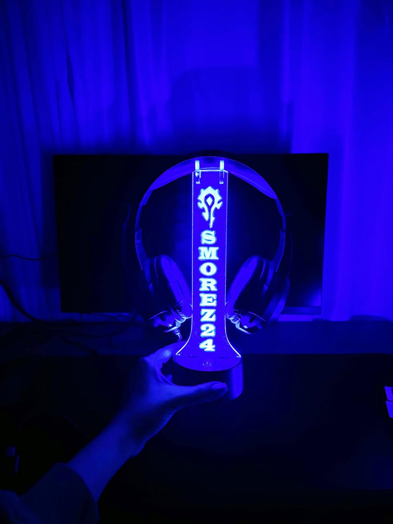 Peut inclure: Un support de casque bleu illumin&eacute; avec le texte "SMORENZ24" et un logo blanc. Le support est con&ccedil;u pour maintenir un casque et est &eacute;clair&eacute; d'une lueur bleue. Le support est tenu par une main.