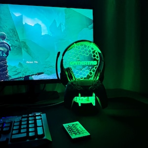 May include: A green gamer headset with a light-up base that says "GAMERTAG" in green. The headset is sitting on a desk with a keyboard and a remote control.