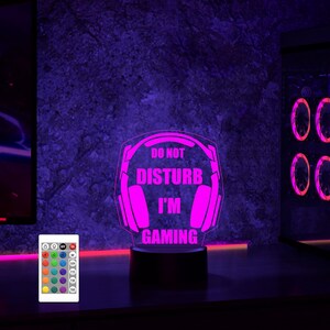 Puede incluir: Una luz nocturna LED rosa con un auricular para juegos y el texto "No molestar, estoy jugando". La luz est&aacute; sobre una base negra y tiene un mando a distancia con m&uacute;ltiples opciones de color.