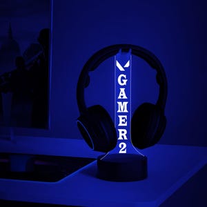 May include: A black gaming headset sits on a blue LED stand. The stand displays the word "GAMER2" in white. The scene is bathed in blue light, creating a modern aesthetic. The setup is designed for gamers.