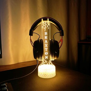 May include: A lit-up headphone stand with the name "BRAXTON" vertically displayed. The stand has a lightning bolt design and holds a pair of black and red headphones. The base and the top of the stand are illuminated with a warm glow.