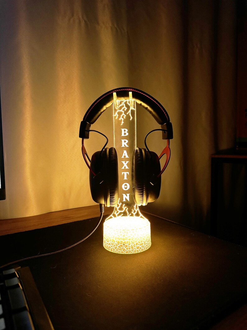 May include: A lit-up headphone stand with the name "BRAXTON" vertically displayed. The stand has a lightning bolt design and holds a pair of black and red headphones. The base and the top of the stand are illuminated with a warm glow.