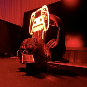 Pu&ograve; includere: Un supporto in acrilico rosso illuminato con un design di controller per videogiochi. Il supporto ha il testo "Gamer 123". Un auricolare da gioco nero e un controller nero sono sul supporto.
