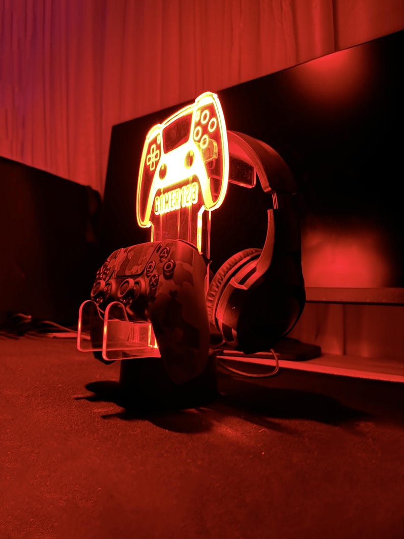 Pu&ograve; includere: Un supporto in acrilico rosso illuminato con un design di controller per videogiochi. Il supporto ha il testo "Gamer 123". Un auricolare da gioco nero e un controller nero sono sul supporto.