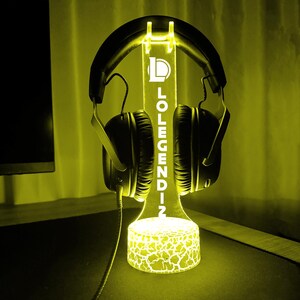 May include: A glowing yellow headphone stand with the text "LOLEGENDZ". The stand is designed to hold headphones and features a cracked pattern on the base. Black headphones are placed on the stand.