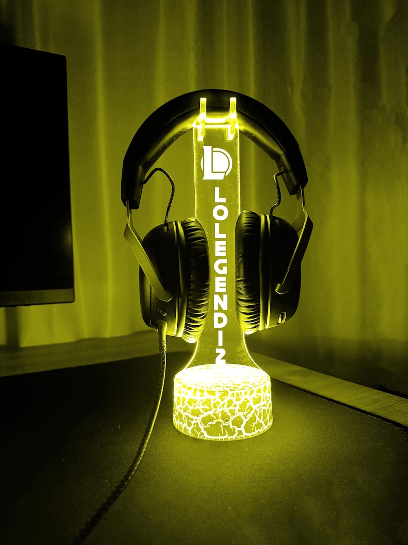 May include: A glowing yellow headphone stand with the text "LOLEGENDZ". The stand is designed to hold headphones and features a cracked pattern on the base. Black headphones are placed on the stand.
