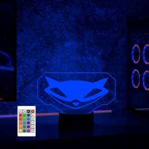 May include: A blue LED night light shaped like the logo for the video game "Sly Cooper". The light is on a black base and has a remote control with multiple color options.