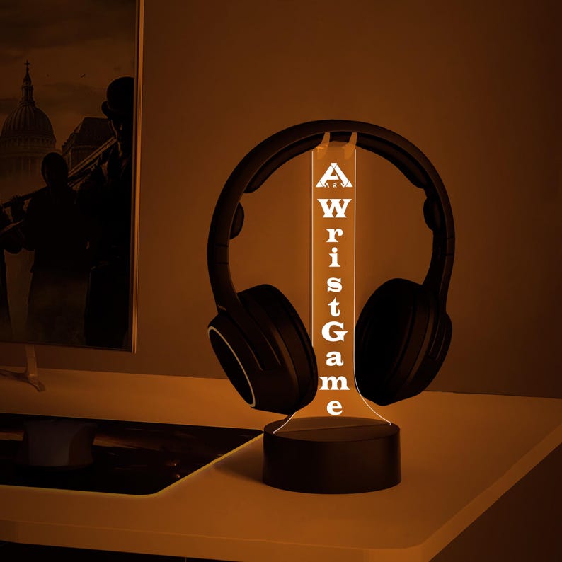 May include: A black and white 3D printed lamp with the word "Wrist Game" printed on it. The lamp is lit up with a warm orange glow and has a pair of black headphones resting on it.