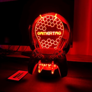 May include: A red illuminated stand with a honeycomb design and the word "GAMERTAG" in yellow. A black video game controller is resting on the stand.