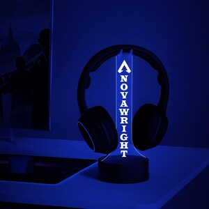 Op de afbeelding: Een witte LED-lamp met het woord "NOVAWRIGHT" verticaal in witte letters gespeld. De lamp staat op een zwarte voet en staat op een bureau naast een zwarte koptelefoon.