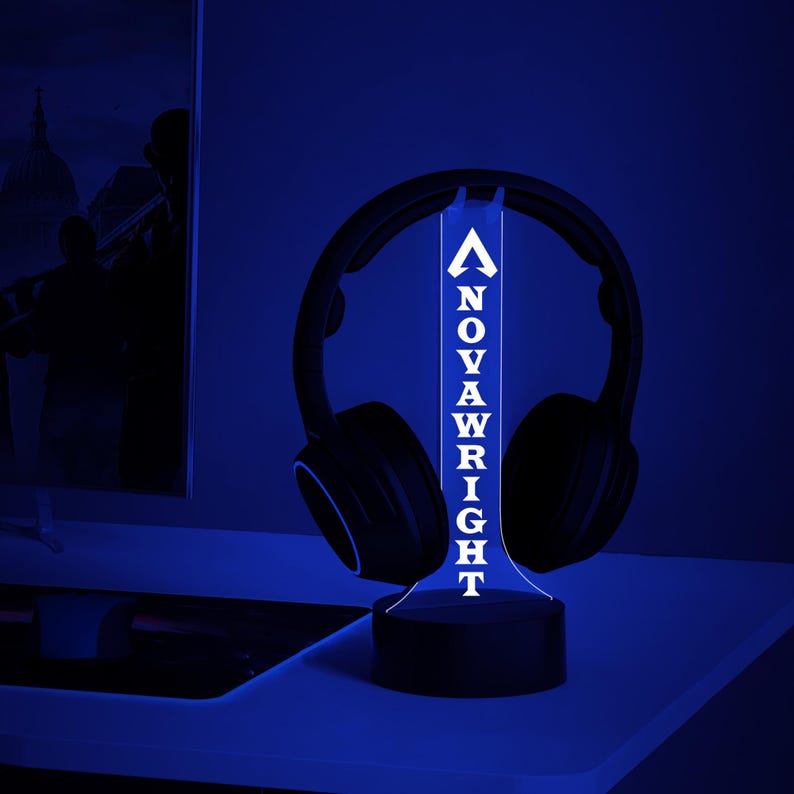 Op de afbeelding: Een witte LED-lamp met het woord "NOVAWRIGHT" verticaal in witte letters gespeld. De lamp staat op een zwarte voet en staat op een bureau naast een zwarte koptelefoon.