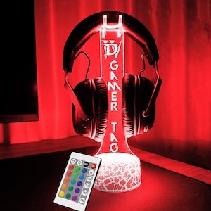 May include: A white LED light stand with a gamer tag design and a remote control. The stand is holding a pair of black headphones. The light is on and is red.