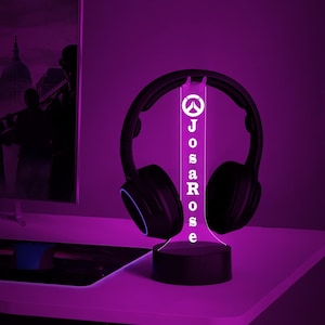 May include: A black and white Overwatch logo with the name "JosaRose" printed below it on a white acrylic base. The base is lit up with a purple light and has a black headphone stand. The headphones are black with a blue accent.