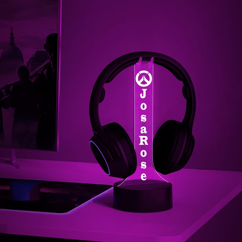 May include: A black and white Overwatch logo with the name "JosaRose" printed below it on a white acrylic base. The base is lit up with a purple light and has a black headphone stand. The headphones are black with a blue accent.