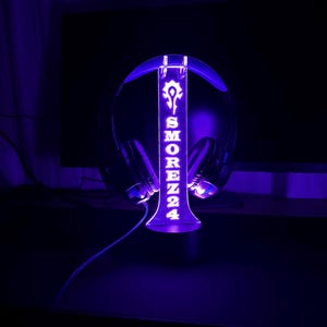 May include: A purple LED light-up headset stand with the name "SMOREZ24" and a symbol on it. The stand is holding a black headset.