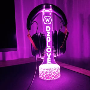 Op de afbeelding: Een paarse LED-lichtstandaard met een wit logo met de tekst "W D2OLOVE" en een zwarte koptelefoon op de standaard.
