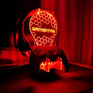 May include: A red illuminated gaming headset stand with a honeycomb design and the word "GAMERTAG" in yellow. The stand also includes a slot for a video game controller.