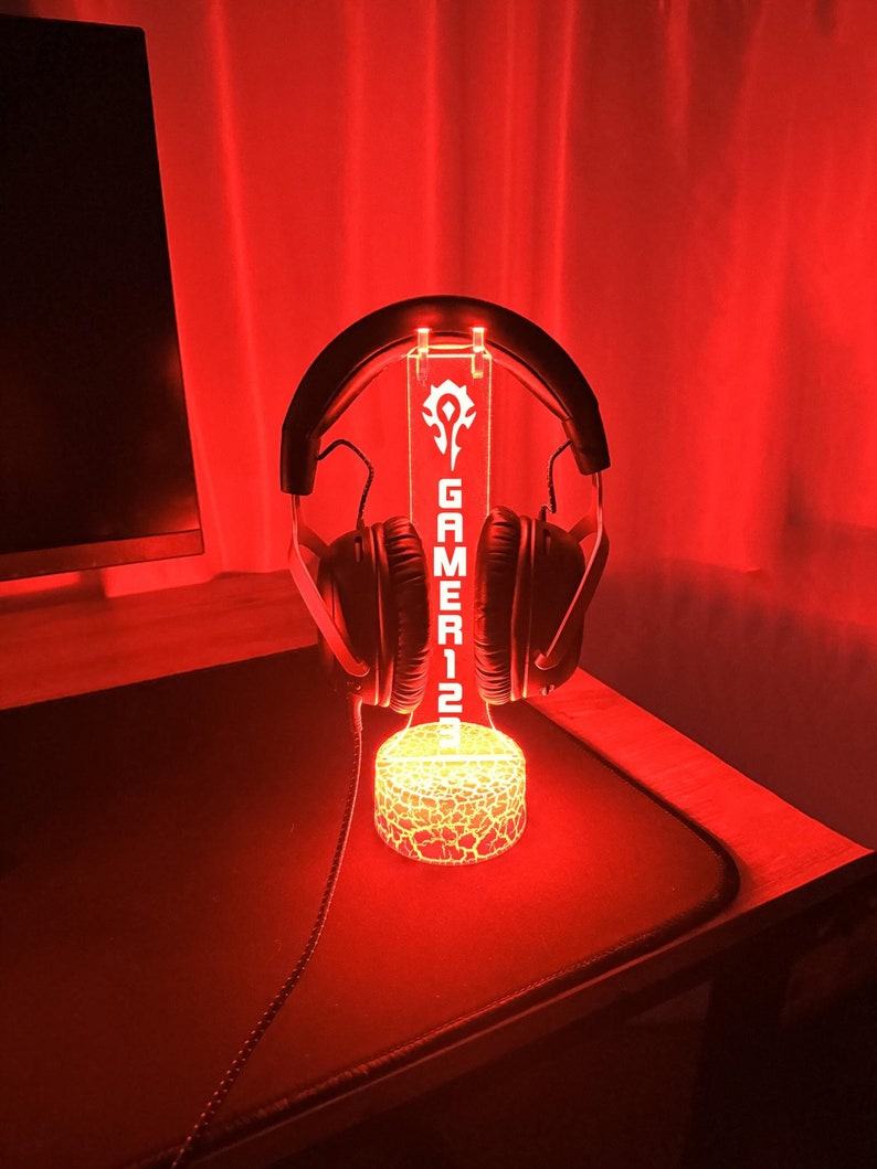 Puede incluir: Una base de luz LED roja con un logotipo de jugador y la palabra "GAMER12" en ella. Un auricular negro descansa sobre la base.