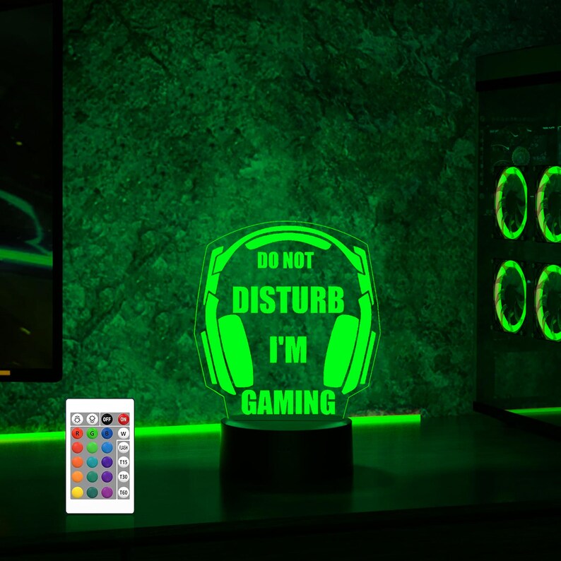 Puede incluir: Luz nocturna LED verde con una base negra y un dise&ntilde;o de auriculares con el texto "No molestar, estoy jugando". La luz se controla mediante un mando a distancia con m&uacute;ltiples opciones de color.