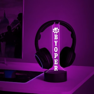 Op de afbeelding: Een witte LED-bureaulamp met een zwarte basis en een transparant acryl paneel met de tekst "BYOPER" en een schedel- en tandwiellogo. De lamp staat op een bureau met een paar zwarte koptelefoons die erop rusten.