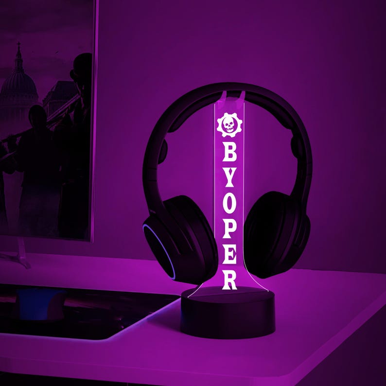 Op de afbeelding: Een witte LED-bureaulamp met een zwarte basis en een transparant acryl paneel met de tekst "BYOPER" en een schedel- en tandwiellogo. De lamp staat op een bureau met een paar zwarte koptelefoons die erop rusten.