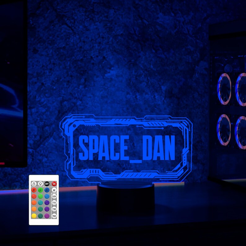 May include: A blue neon sign with the text "SPACE_DAN" in a futuristic font. The sign is on a black base and is illuminated with a blue light. A remote control with multiple color options is in the foreground.