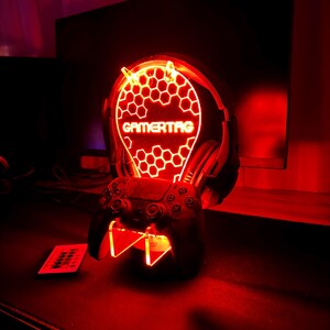 May include: A red illuminated gaming headset stand with a honeycomb design and the word "GAMERTAG" in the center. A black video game controller is attached to the stand.
