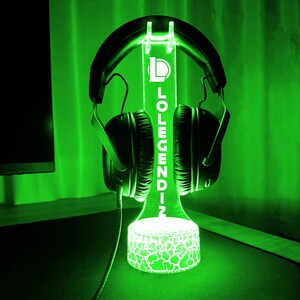May include: A green LED headphone stand with the text "LOLEGENDZ" vertically displayed. The stand has a cracked pattern at the base and holds a pair of black headphones. The background is a solid green.