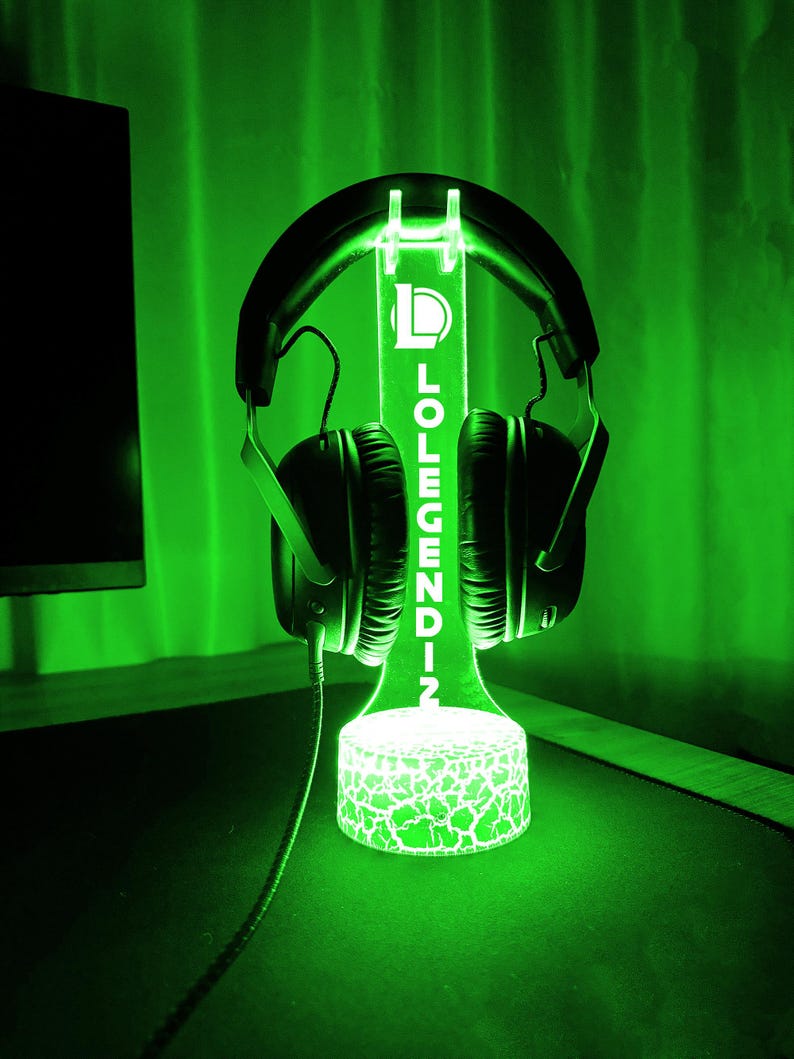 May include: A green LED headphone stand with the text "LOLEGENDZ" vertically displayed. The stand has a cracked pattern at the base and holds a pair of black headphones. The background is a solid green.