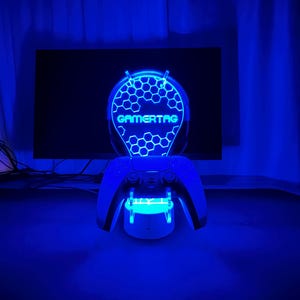 May include: A blue LED light with a honeycomb design and the text "GAMERTAG" in white. The light is mounted on a stand that holds two black video game controllers.