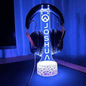 May include: A blue LED light-up stand with a white Overwatch logo and the name "JOSHUA" on it. A black and orange headset is resting on the stand.